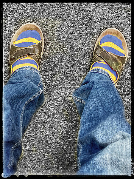 Person med blåjeans och bruna sandaler över gula och blå randiga strumpor på grått golv.