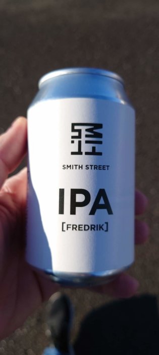 Ölburk med etikett "SMITH STREET IPA [FREDRIK]" hålls i handen, utomhus med suddig bakgrund.