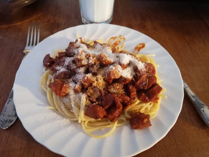 Tallrik med spagetti och skivad korv toppad med riven parmesan, serverad på ett mörkt träbord.
