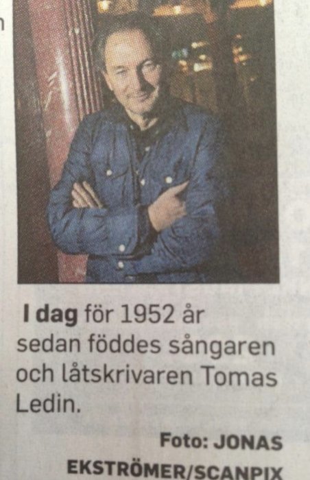 Man i jeansjacka med armarna korsade framför tidningsartikel med text.