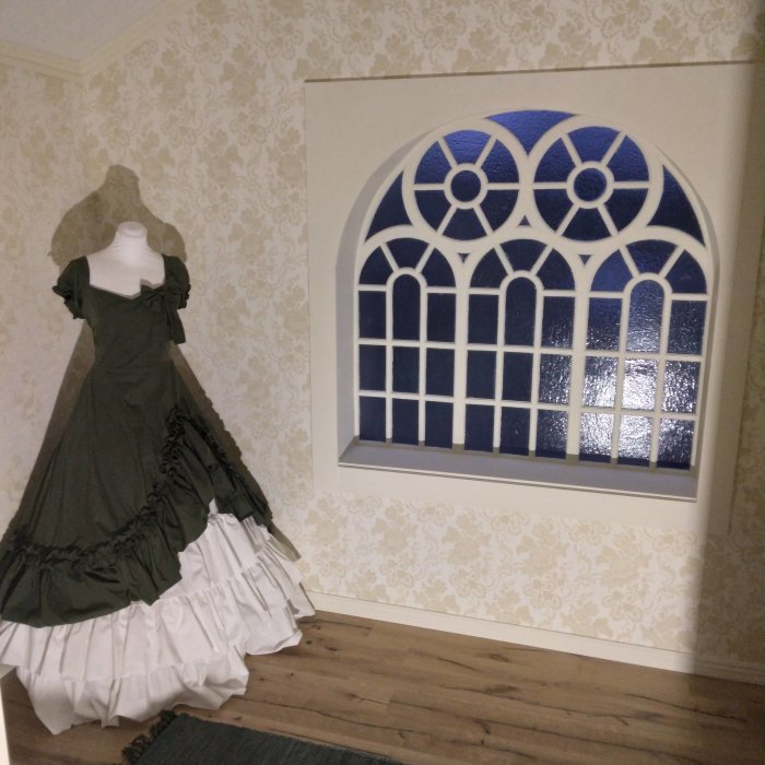 En viktoriansk stil klänning på en provdocka bredvid ett fönster med glasmålning i en rum med tapetserade väggar.