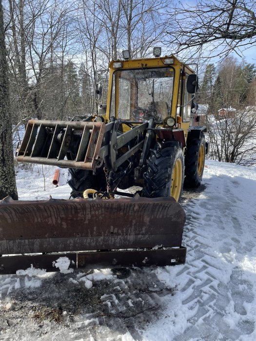 Gul traktor med snöplog framför på en snöig väg, omgiven av träd och snödrivor.