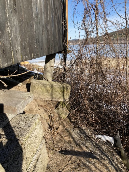 Grund av ett uthus på en sandig slänt med erosion, synlig ny hängränna och snörester i bakgrunden.