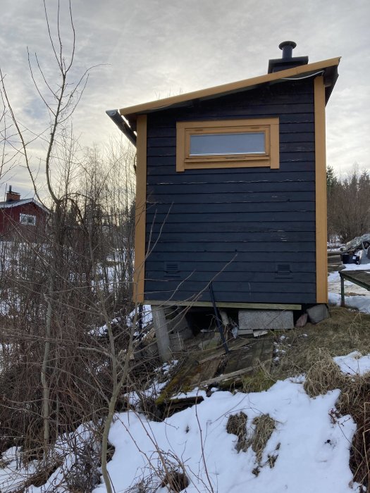 Lutande litet blått uthus med ny hängränna på en sandig slänt med snöfläckar och viss vegetation.