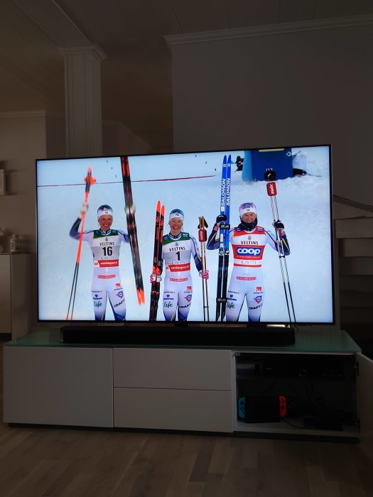 TV-skärm visar tre skidåkare med pjäxor och skidor som firar på en vinnarpall.