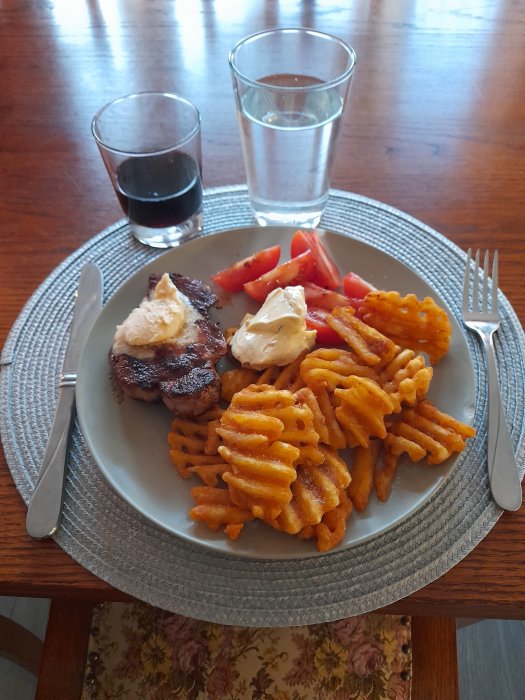 Fläskkotlett med vitlökssmör, waffled pommes frites och tomat på en tallrik, med vattenglas och vin.