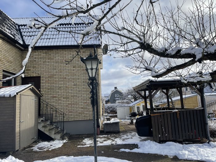 Ett snötäckt bakgård med hus, lyktpelare och pergola under en klar vinterdag.