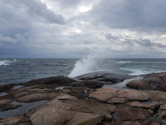 Molnig himmel ovanför en havsvik med vågor som slår mot klipporna och skapar stänk.