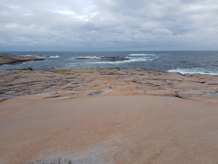 Sundslipt klippa vid havet med vågor som bryter mot strandkanten under molnig himmel.