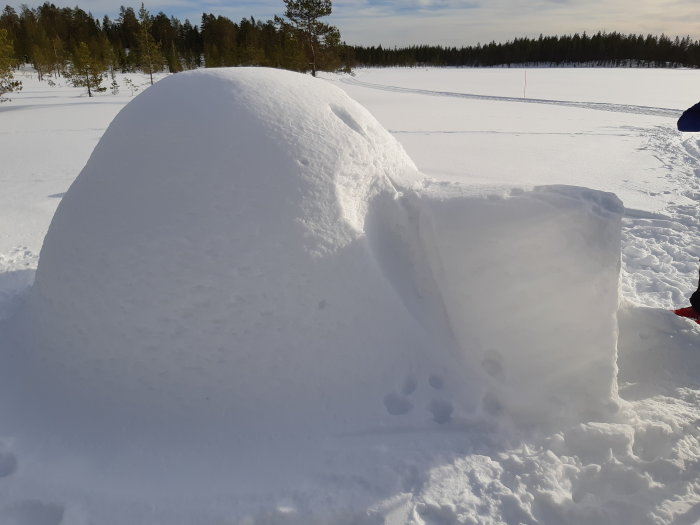 Igloo av snö med entréblockerad av stor snöbit, i soligt vinterlandskap.