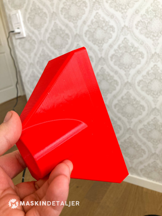 Hand håller en röd, 3D-utskriven adapter för dammuppsamlare mot en tapetserad vägg.