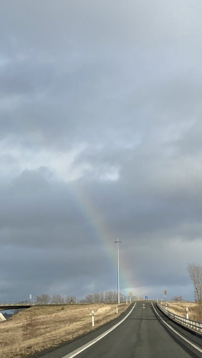 Väg som leder mot en regnbåge i ett öppet landskap med delvis molnig himmel.