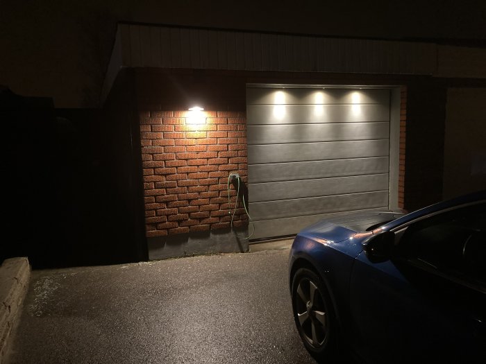 Kraftfulla lampor monterade på garagevägg lyser upp garageport och uppfart med blå bil parkerad framför.