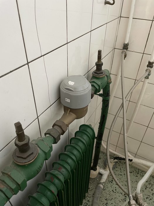 Äldre stängventil och vattenmätare intill en grön radiator och vit kaklad vägg, möjligt behov av reparation.