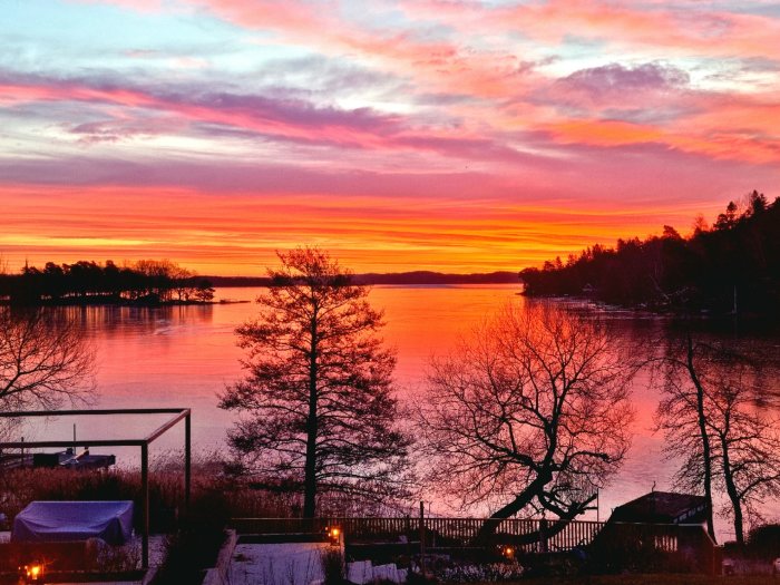 Landskap vid sjö med levande ljus vid solnedgång, färgglad himmel och träd i silhuett.