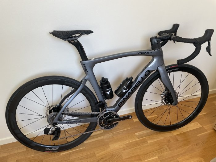 Grå Pinarello Dogma F12 racercykel stående mot en vit vägg på ett trägolv.