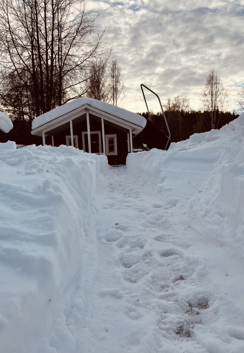 Vit snötäckt stuga med uppskottad stig och höga snöväggar mot molnig himmel.