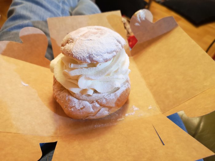 Semla med fluffig grädde på ett brunt papper, ben person i bakgrunden.