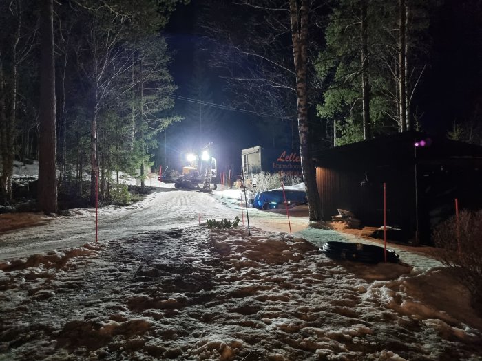 Grävmaskin som arbetar på natten för kabelutsättning och borrning i en snöig trädgård med markeringsstolpar.