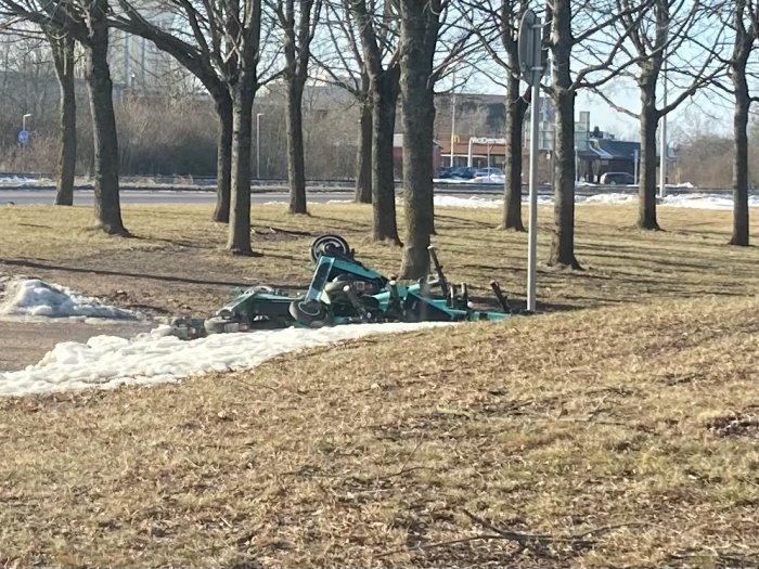 Grönt redskap för markarbete omkullvält på en gräsmatta med träd i bakgrunden.