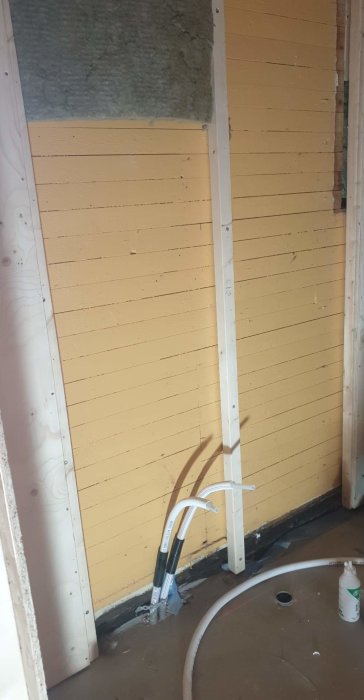 Ofärdigt badrum med isolering och gula väggpaneler, rörfixaturer som sticker ut ur väggen och justerad elinstallation.