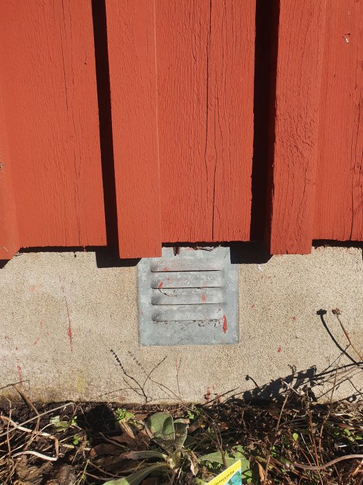 Ventilationsgaller i grått på en husgrund med röda bräder ovanför och vegetation nedanför.