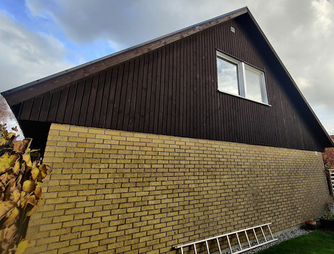 Gavel på ett hus med brun träpanel och ett stort fönster, samt en gul tegelgrund.