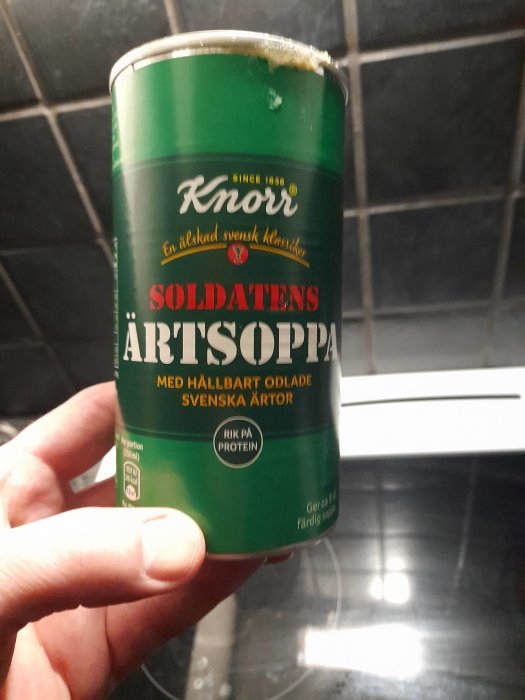 Hand håller en burk av Soldatens Ärtsoppa från Knorr mot en kaklad köksvägg.