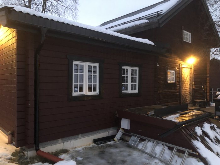 Rödmålad timmerstuga med vita fönster under skymningen, snö på taket, byggmaterial synligt framför huset.