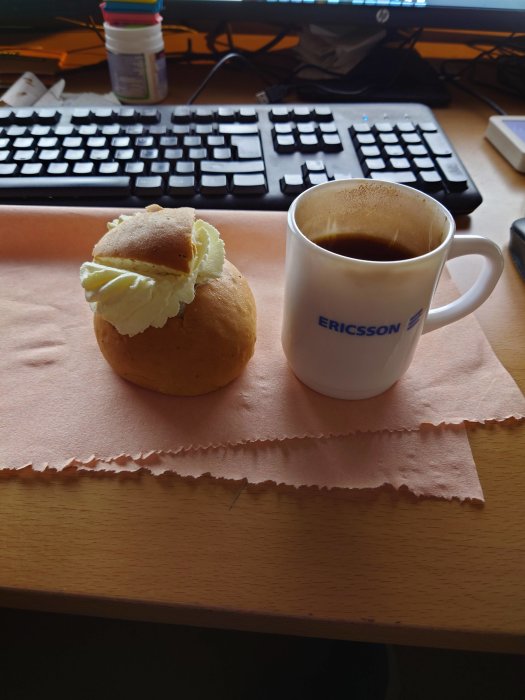 Semla med vispgrädde och kaffe i Ericsson-mugg på skrivbord med tangentbord i bakgrunden.