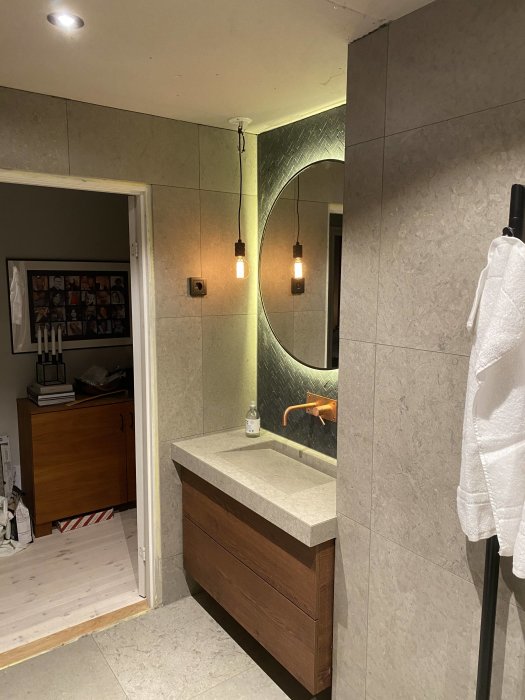 Halvfärdigt badrum med rund spegel, upphängda lampor och träkommod.
