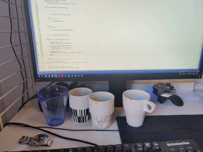 Fyra använda kaffemuggar på skrivbord framför datorskärm med kod, tangentbord och spelkontroll.