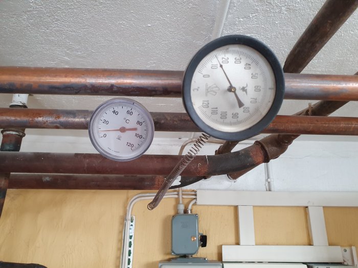 Två runda manometrar på kopparledningar, en för temperatur och en för tryck, i ett värmepumpssystem.