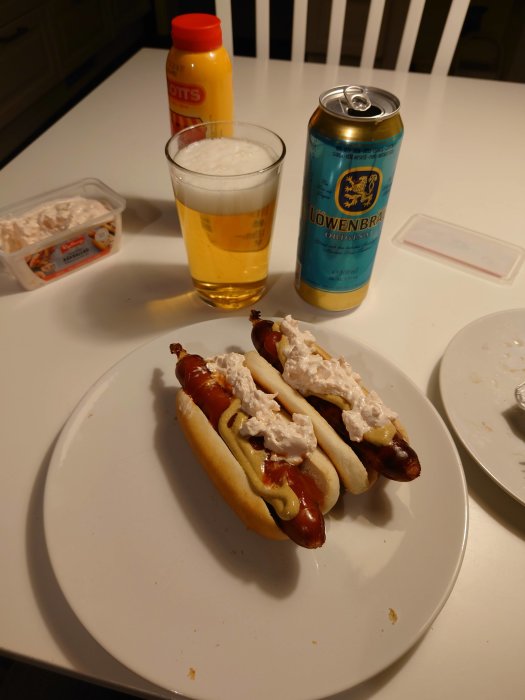 Två korv med bröd med senap och toppad med räksallad, ett glas öl och en burk Löwenbräu på ett vitt köksbord.