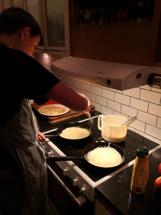 Barn steker pannkakor i kök med spis och stekpannor, smet i kanna.