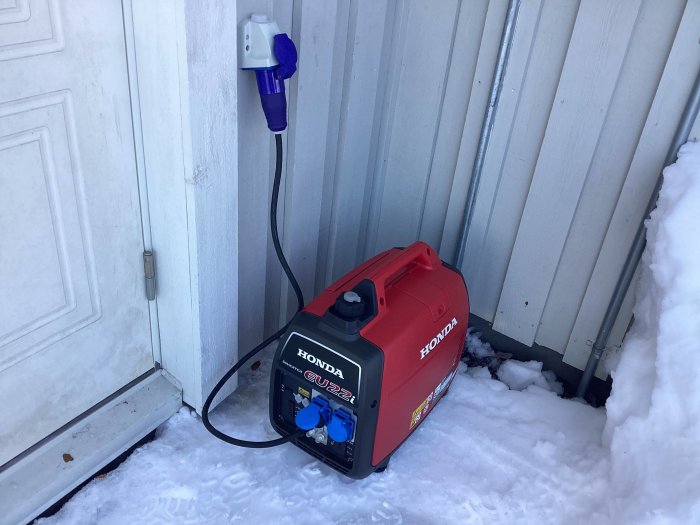 Röd bärbar Honda-generator kopplad till strömuttag på husfasad med snö omkring.