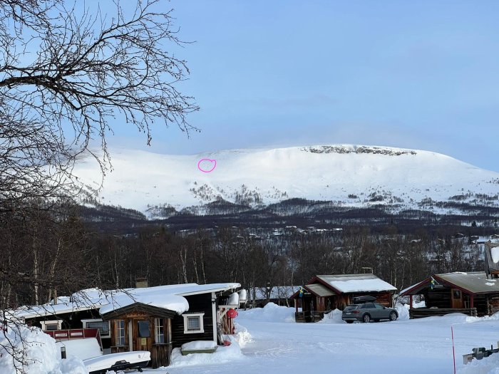 Vinterlandskap med snöklädda fjäll och stugor, trädgren i förgrunden.