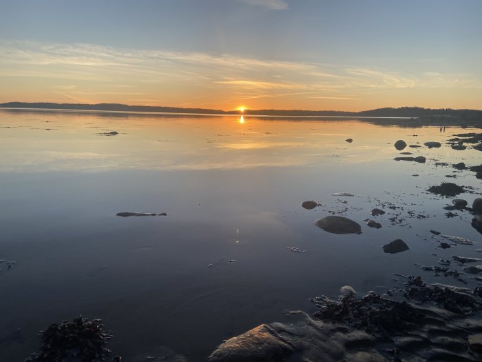 Solnedgång vid havet med spegling på vatten och stenar i förgrunden.