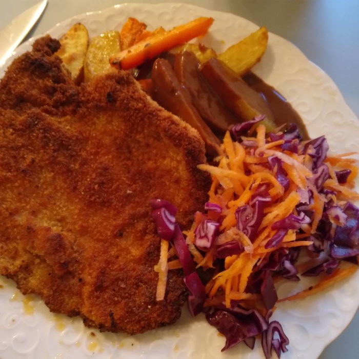 Panerad schnitzel med ugnsbakade potatisklyftor, morot, rödvinssås och råkostsallad på en vit tallrik.