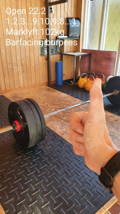 Tummen upp framför hemmagym med skivstång, vikter och kettlebells, planeringsanteckningar på väggen.