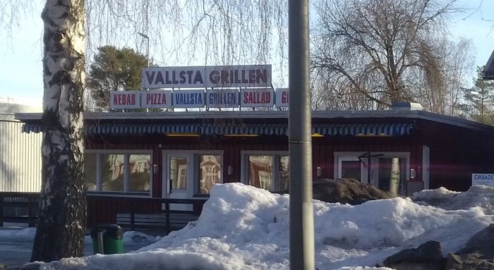 Grillrestaurang med skylt "Vallsta Grillen" omgiven av snö och en björkträd till vänster.