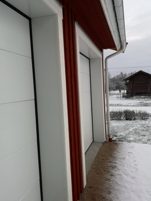 Hörn av byggnad med plåtsalning och hörnlist synliga nära garageport och snötäckt landskap i bakgrunden.