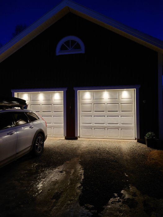 Mörk fasad med två upplysta garageportar på kvällen, bil parkerad på sidan, och reflekterande ljus på marken.