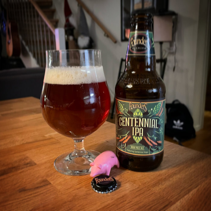 Ölglas fyllt med rödbrun öl bredvid en flaska Founders Centennial IPA och en liten rosa figur på ett bord.