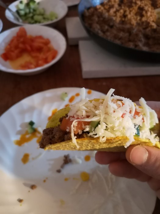 Hand håller en taco med köttfärs, ost och tomat, suddiga skålar med ingredienser i bakgrunden.