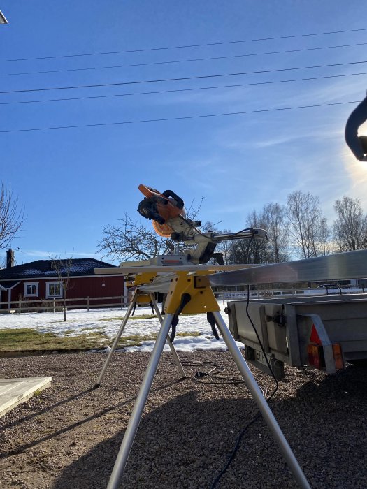 Sågstation utomhus med kap- och geringssåg på stativ framför släpvagn och snöfläckat gräs.