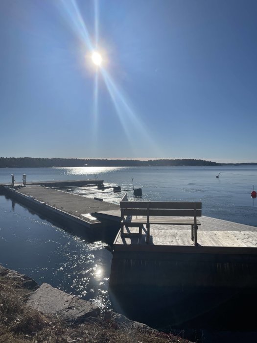 Solig vy över en brygga med sjö och skog i bakgrunden, solreflektioner på vattnet.