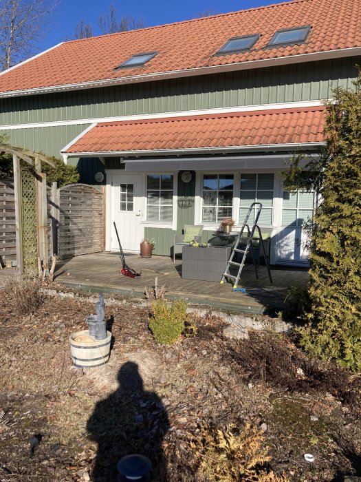 Ett hus med röd takpannor och grön träfasad. På verandan finns en stege, på gräset står en skottkärra.