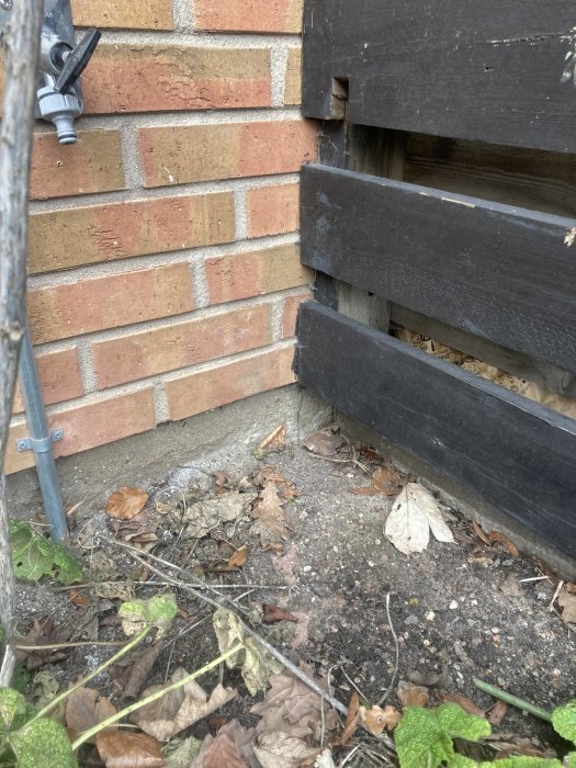 En gammal sliten spricka vid gränsen mellan två radhus under ett svart plank, vid en tegelvägg och betonggrund.