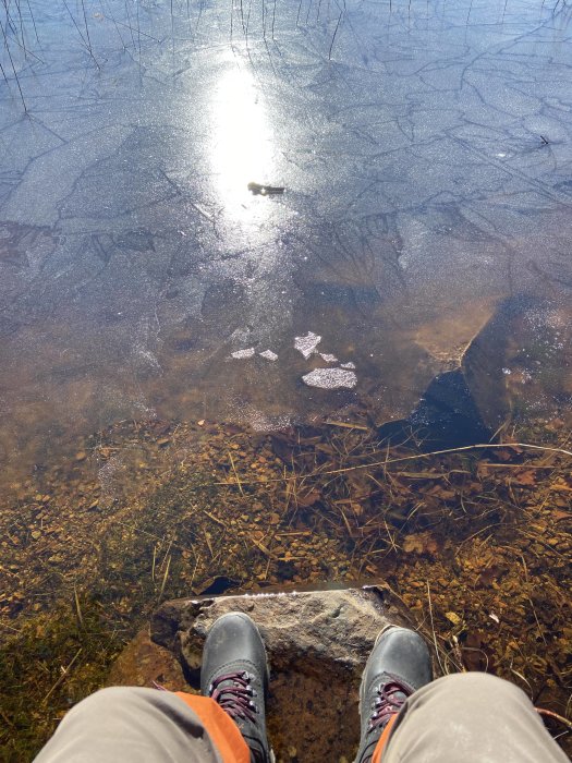 Person som sitter vid en isbelagd sjö med solreflektion, ser ner på sina kängor och klippig strand.